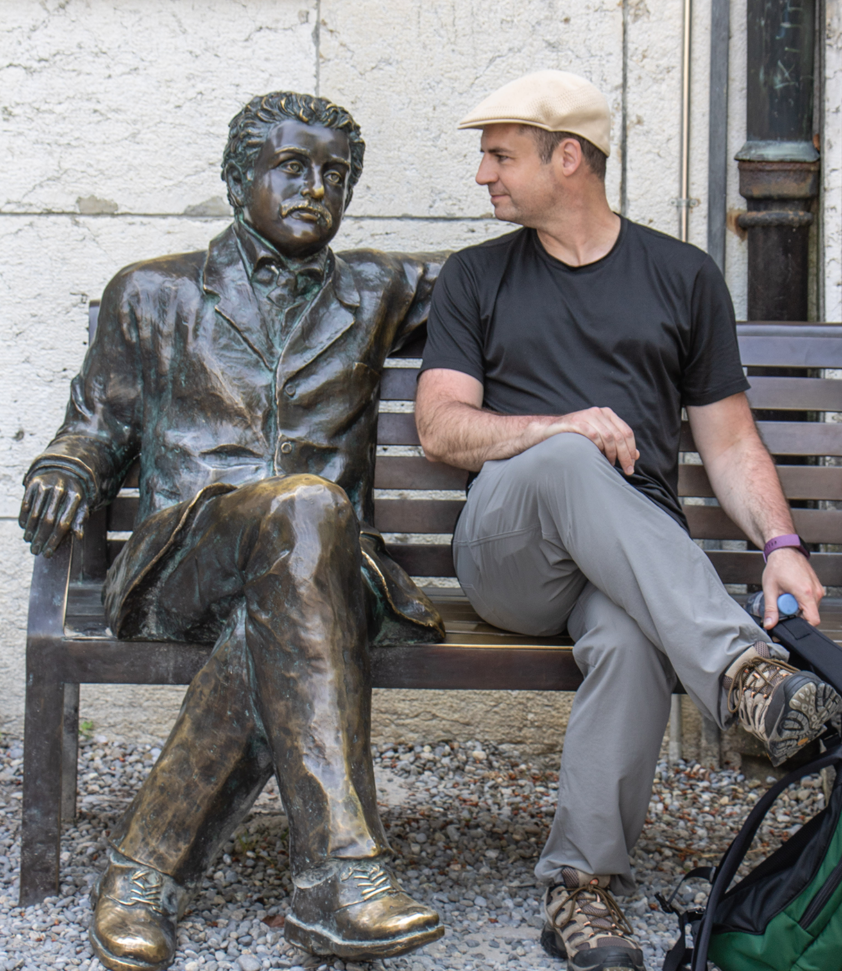Jason Roth, wearing a black t-shirt and beige Kangol cap, sits on a bench next to a bronze statue of Albert Einstein in formal attire and appears to have a conversation with the statue