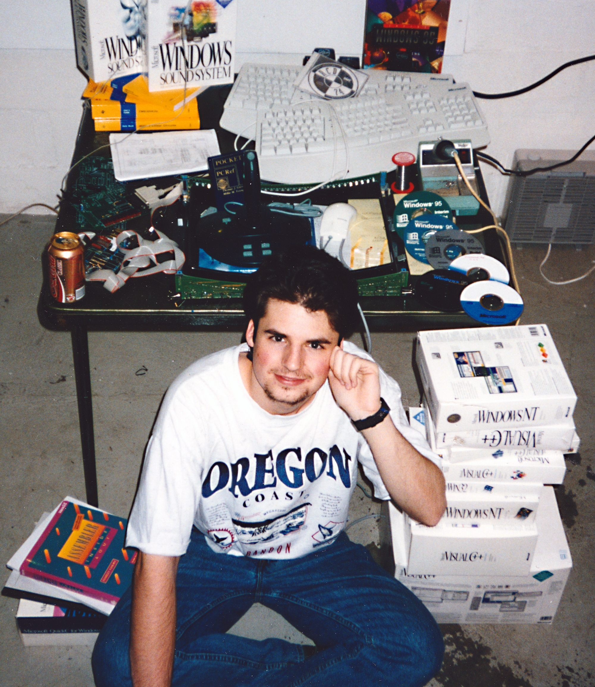 In his high school years, Jason Roth, wearing a white Oregon Coast t-shirt, sits in front of a cluttered table with a computer keyboard and stacks of CDs and software boxes including Windows 95, smiling at the camera