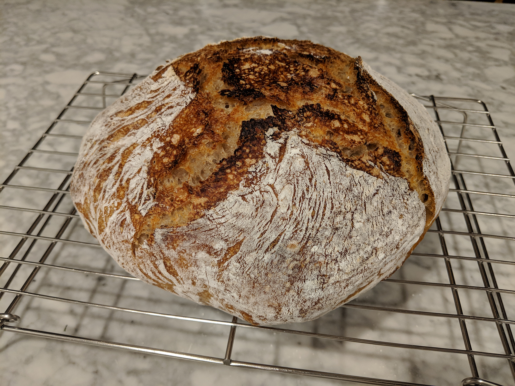 A rustic artisanal sourdough bread loaf with a golden-brown crust and flour dusting sits cooling on a wire rack, showing the characteristic scoring patterns and texture of handmade bread